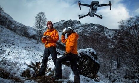 Dos rescatistas con ropa naranja observan un dron sobre un paisaje nevado